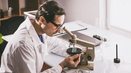 Naklejka na ściany i meble An adult male Caucasian scientist in glasses and a white coat works with a sample under study on a microscope. Color correction