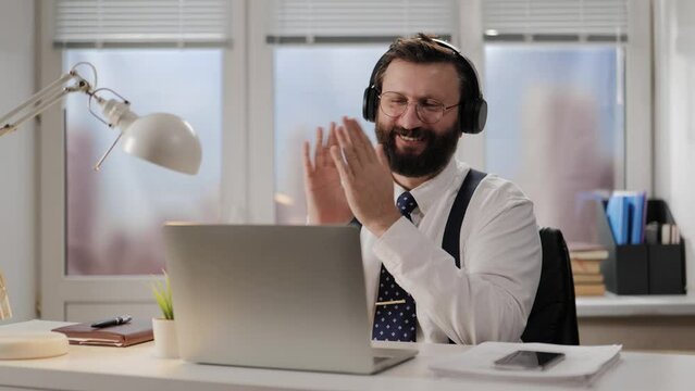 Success, satisfied businessman applauds. Positive man in headphones, glasses and tie applauds on webcam. Office worker applauding at online conference