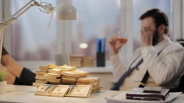 Success, earnings, good deal. Successful businessman with glass of whiskey in his hand drinks alcohol and celebrates success. There is large pile of money in foreground
