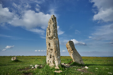 Two ancient, weathered standing stones in a grassy field under a blue sky with scattered clouds