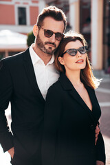 Beautiful fashion woman and her handsome elegant boyfriend in black suit. Sexy brunette model in jacket. Fashionable confident couple posing in street. Brutal man and female outdoors. In sunglasses
