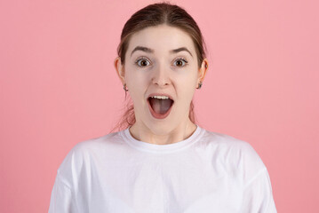 Close-up portrait of an attractive Caucasian young brunette woman in a casual white t-shirt who is shocked by something isolated on a pink studio background.