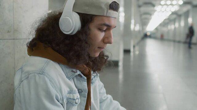 Side tilt footage of 20 yo man with puffy curly hair listening to music in headset and chatting on mobile while waiting for underground train in long hallway