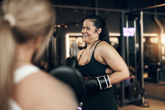 Young woman wearing boxing gloves and a tank top laughing while practicing punching with a female partner holding pads during a self defence workout at the gym