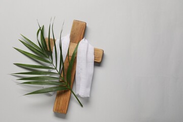 Wooden cross, white cloth and palm leaf on light grey background, top view with space for text. Easter attributes