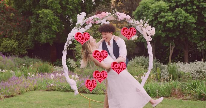 Animation of red love you hearts over happy diverse couple embracing in garden on wedding day