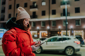 Side view of pretty young woman wearing hat and winter jacket calling taxi using smartphone app. Frozen smiling female calling mobile phone waiting taxi in evening cold street.