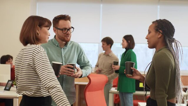 Diverse team of relaxed workmates chatting, enjoy conversation during coffee break at workplace. Friendly male and female colleagues having informal talk, drink tea, resting before or after workday.