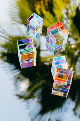 Glass geometric prism cubes with light diffraction of rainbow spectrum colors on blue sky and palm...