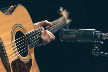 Male musician playing acoustic guitar behind microphone in recording studio.