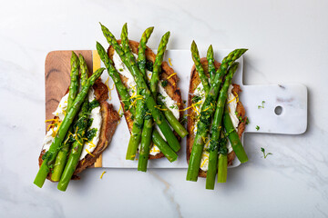 Grilled toasts with sourdough bread with green asparagus, ricotta, lemon zest and herb oil on marble white board.