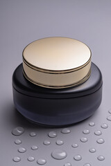 Luxury face cream jar with golden lid and matte glass on grey background with waterdrops. Packaging...