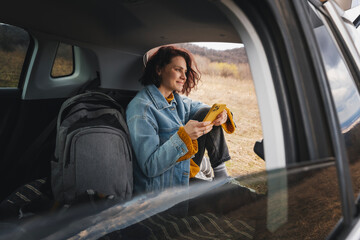Young happy woman sitting in an open car trunk taking pictures with a camera. Traveling by car, communication in travel concept - 746381019