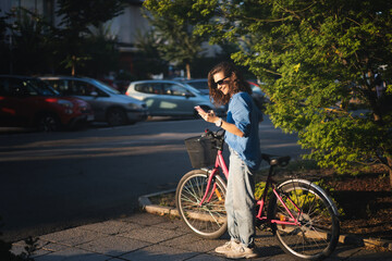 A young Caucasian woman using a smartphone while standing next to her bicycle in the summer city street. - 746381897