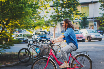 A young happy Caucasian woman rides a bicycle to a public bicycle parking lot in a summer city - 746384653