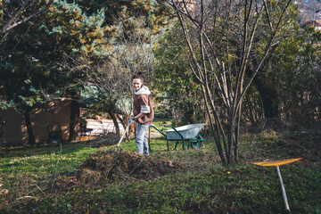 A young woman removes dry grass and cleans the soil in the spring garden of a country house. Spring work in the garden, preparing land for planting - 746385259