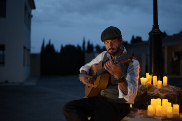 Guitarist in waistcoat and beret playing the Spanish guitar by candlelight in the village square.
