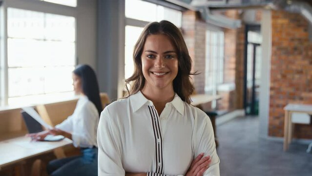 Portrait of smiling young businesswoman working in modern open plan office turning to look at camera and folding arms - shot in slow motion