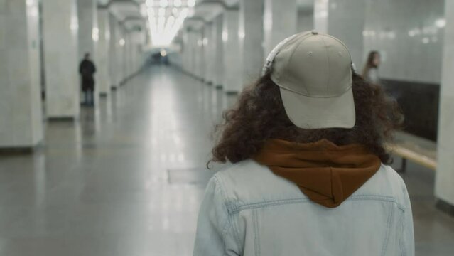 Tilt tracking footage of young handsome Caucasian curly-haired skateboarder with bright board in hand walking along lobby in tiled metro underpass and turning around