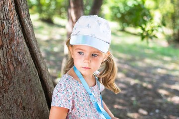 Little girl in a floral dress and a baseball cap standing near a tree in the park