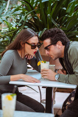 Young stylish attractive couple wearing sunglasses drink with the straw in the same glass a cocktail sitting in a terrace outdoors. Human relationships, beautiful people drinking alcoholic drinks.