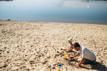 Father and 3 years son on beach building sand castle with plastic toys . single father solo parent. Involved parenting. Relaxing and playing outdoors with family