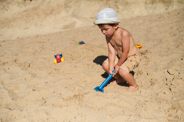 A 3-year-old child builds a sand castle on the beach. Games on the beach on a hot summer day. A boy plays with a stove and makes little beads, pours sand into a bucket with a plastic scoop
