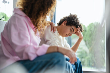 Upset teenage boy sitting on sofa while his mother looking at him
