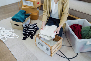 Asian young woman packing clothes at home, putting on stuff into donate box with second hand clothes