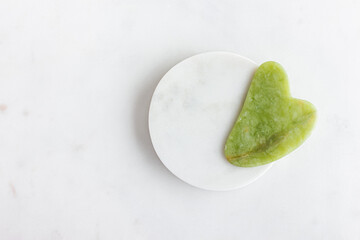 Green jade gua sha massager for face on white marble.