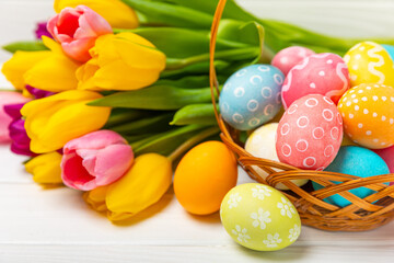 Fototapeta na wymiar Easter basket filled with colorful eggs and a bouquet of tulips on a textured wooden table. Easter celebration concept. Colorful easter handmade decorated Easter eggs. Place for text. Copy space