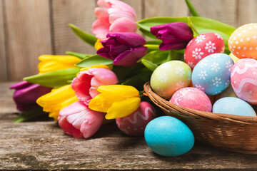 Easter basket filled with colorful eggs and a bouquet of tulips on a textured wooden table. Easter...