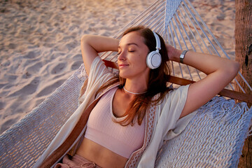 Young blissful calm woman wearing wireless headphones with closed eyes and hands behind head lie in a hammock and enjoy relaxing music