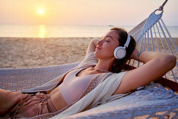 Young blissful calm woman wearing wireless headphones with closed eyes and hands behind head lie in a hammock and enjoy relaxing music