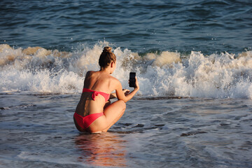 Girl in bikini taking photo on smartphone camera of surf waves on sea beach