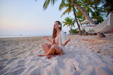 Young blissful calm woman wearing white wireless headphones with closed eyes and lotus pose hands meditates on the sand beach and enjoy relaxing music, mind care