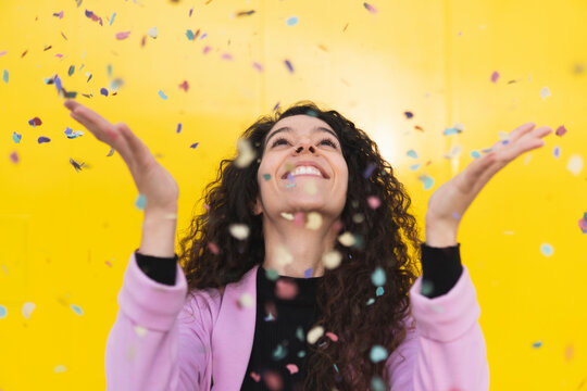 Cheerful woman playing with confetti against yellow background