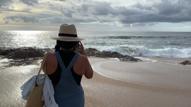 Hispanic dark haired woman taking photo of sunset at beach as tide rolls in