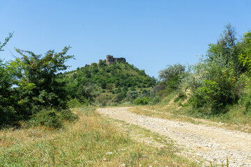 Fototapeta premium A dirt road leads to the ruins of medieval fortress on the hill. Bushes and fields around. Blue bright sky.