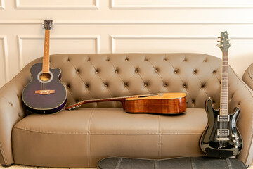 Three guitars set on a brown leather sofa in the living room. A black electric guitar, a brown acoustic and a purple classical guitar. Indoors at home with moldings on a grey wall in the background.