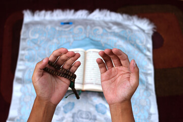 Indonesian man opening his palms in prayer after reciting the Quran on a prayer mat during Ramadan...