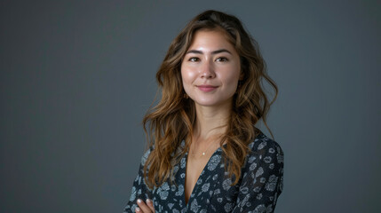 Obraz premium Portrait of a young woman with long wavy hair, wearing a patterned blouse, standing confidently against a neutral grey background.