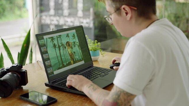 Transgender photographer edits photos on laptop in plant-filled coworking space. Tattooed digital nomad retouches images, embodies diversity, inclusivity in creative industry. Slow motion.
