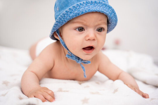 Horizontal  photograph of a baby  with his mouth open and wearing a hat lying on a blanket.