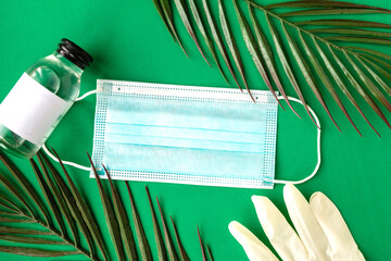 Face medical mask, sanitizer and tropical leafs on green background minimal. Coronavirus travel....