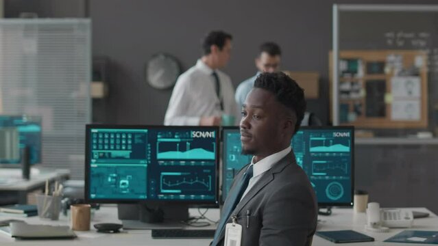 Medium slowmo portrait of young African American male cybersecurity officer turning to camera on office chair and posing for camera, sitting in front of multiple computer displays at office desk