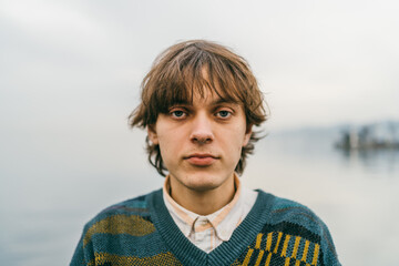 Young male with contemplative gaze, wearing sweater, against blurred lakeside backdrop.