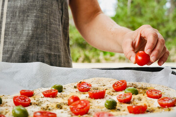 Woman preparing traditional Italian focaccia puts cherry tomatoes on top of the dough. Cooking outside in the garden with fresh ingredients.
