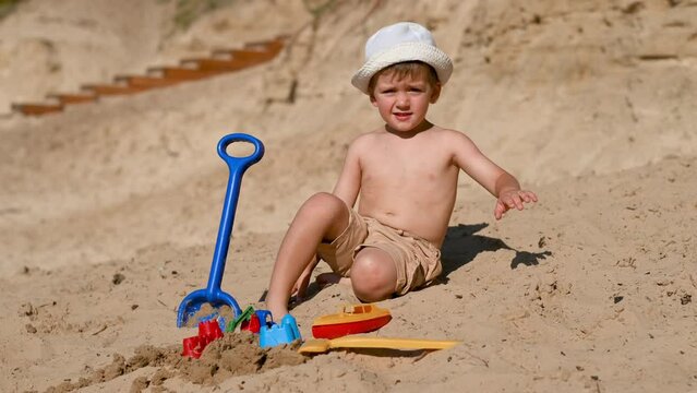 A 3-year-old child builds a sand castle on the beach. Games on the beach on a hot summer day. A boy plays with a stove and makes little beads, pours sand into a bucket with a plastic scoop