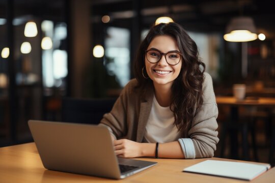 Cheerful woman in casual clothes and glasses sitting at table with laptop and looking at camera while working